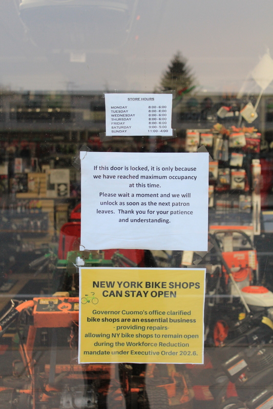 Three signs on a window.
Top sign is store hours.
Middle sign says "If this door is locked, it is only because we have reached maximum occupancy at this time. Please wait a moment and we will unlock as soon as the next patron leaves. Thank you for your understanding and patience." 
Bottom sign says "New York bike shops can stay open. Governor Cuomo's office clarified bike shops are an essential business-providing repairs-allowing NY bike shops to remain open during the Workforce Reduction mandate under Executive Order 202.6."