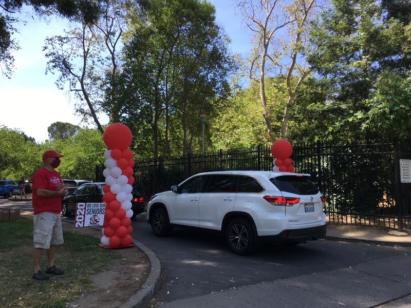 A black sedan followed by a white SUV driving along a road through two pillars of red and white balloons. Next to the balloons is a sign that says "2020 WE LOVE OUR SENIORS"