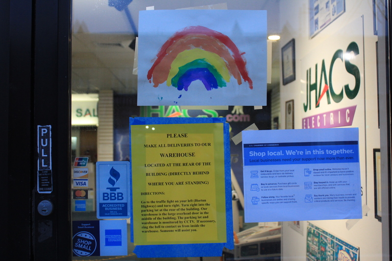 A window with a couple of signs taped to it. There is a sign to shop local on the right, directions to make deliveries to the facilities warehouse on the left, and a rainbow painting that sits above both signs. Through the window, the company JHACS Electric can be seen.