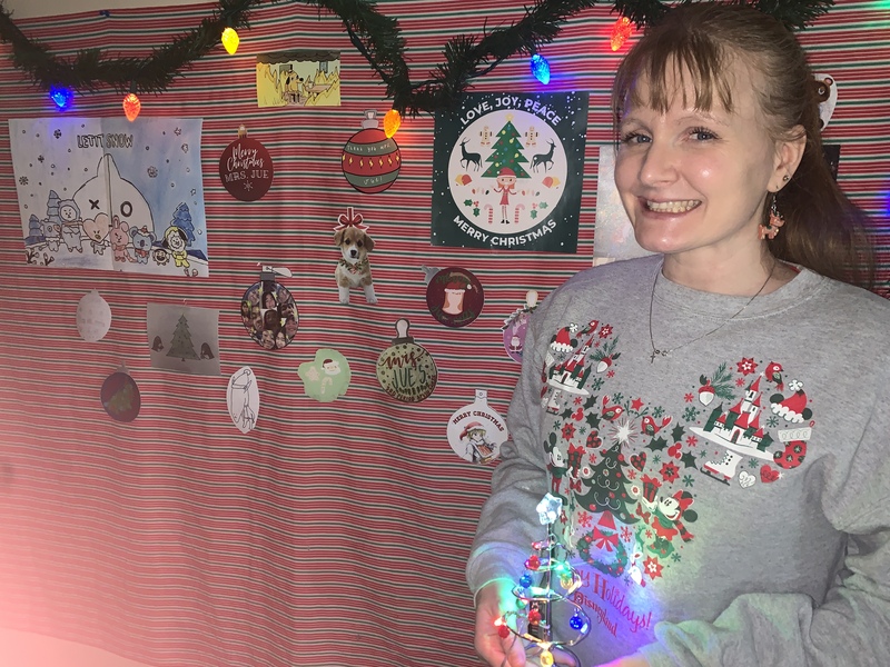A woman in a Christmas sweatshirt, holding a small ornament, in front of a wall decorated for Christmas.