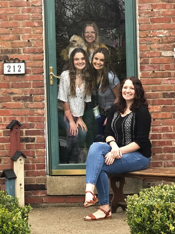 This is a picture which shows a woman sitting on a bench, posing for a picture with three other women who are behind a glass door. All are smiling, and a small dog is being held by one of the women behind the glass door. 