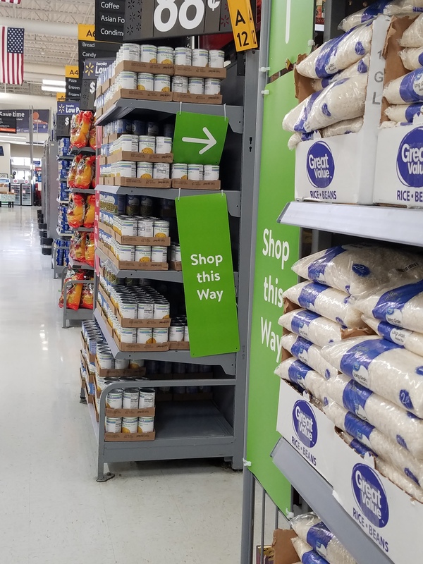 This is a picture taken of an aisle in a store, where a green sign is posted at the entrance reading "Shop this way". 