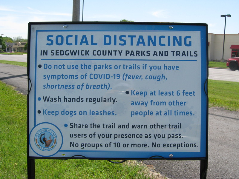 Blue and white sign by Sedgwick County, Kansas with text saying, "SOCIAL DISTANCING IN SEDGWICK COUNTY PARKS AND TRAILS: Do not use the parks or trails if you have symptoms of COVID-19 (fever, cough, shortness of breath).; Wash hands regularly.; Keep dogs on leashes.; Keep at least 6 feet away from other people at all times.; Share the trail and warn other trail users of your presence as you pass. No groups of 10 or more. No exceptions."