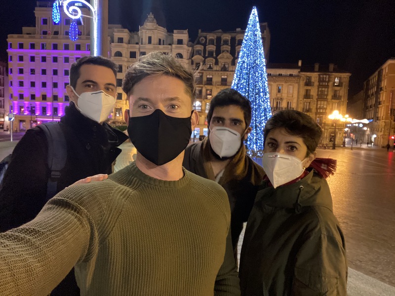 This is a picture of three men and one woman wearing face masks while posing in front of a brightly lit  Christmas tree. 