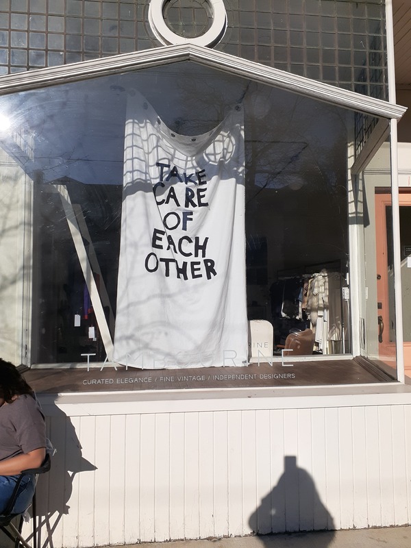 This is a picture taken of a storefront window, were a banner is hung reading "Take care of each other". 