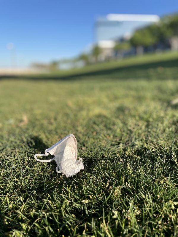 This is a picture of a face mask that has been disposed of in a grassy area. The rest of the picture is out of focus, but a building and trees can be barely seen blurred in the background of the shot. 