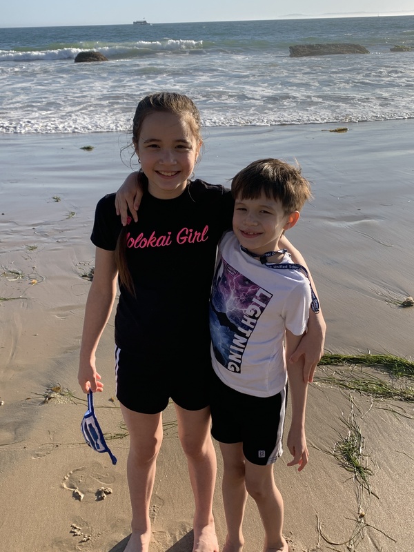 This is a picture of a young boy and girl at the beach wearing shorts and t-shirts, hugging one another to pose for a photo.