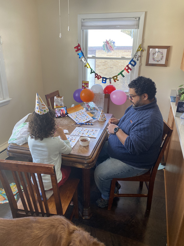 This is a picture of a man and his daughter sitting at a table. They are hearing birthday party hats, and a sign reading "happy birthday" hangs from a window in the background. 