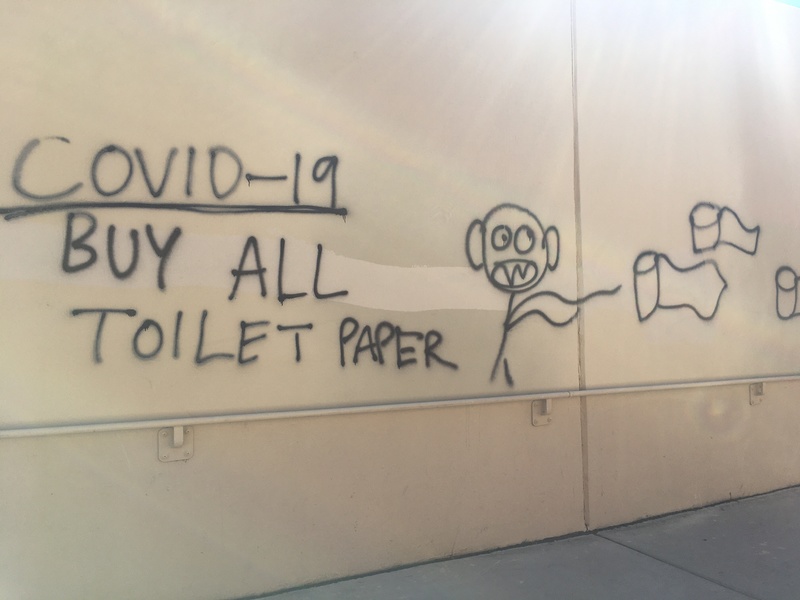 Black graffiti of a wall that says: COVID-19 BUY ALL THE TOILET PAPER. It shows a stick figure running after rolls of toilet paper. 