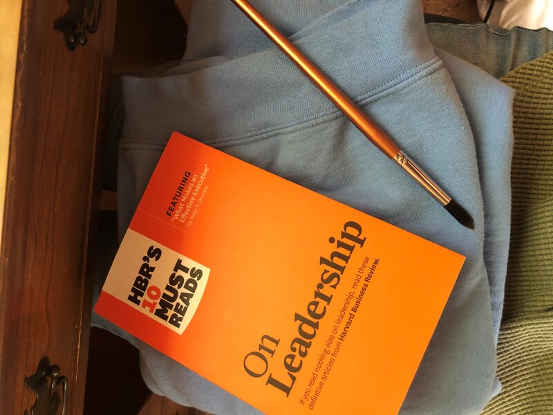An orange book titled "On Leadership" laying on a blue sweatshirt with a paintbrush. 