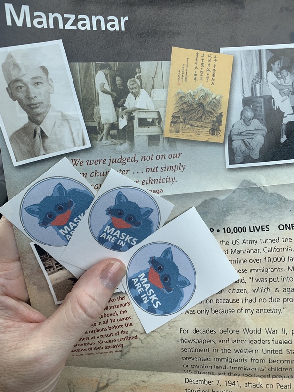 This is a picture taken of stickers promoting the wearing of masks. A program relating to Manzanar Internment Camp sits below it. 