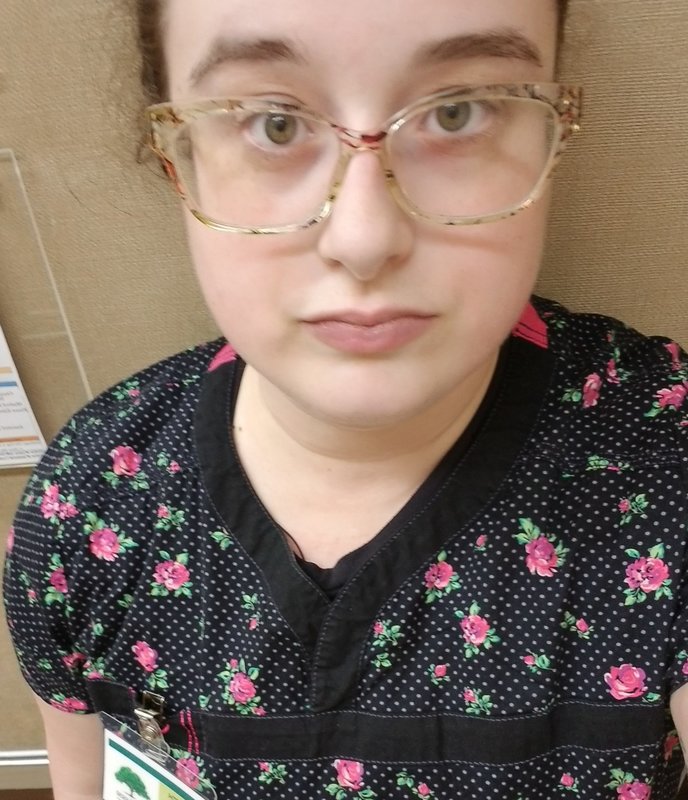 A selfie that is taken by a healthcare worker wearing black and pink scrubs. The person is wearing clear frame glasses. 