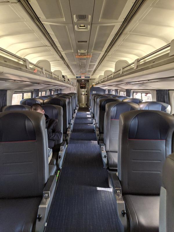 Inside of an Amtrak cabin that only has one person sitting in a seat off to the left. The rest of the cabin is empty. The isle has blue carpet to match the blue headrests of the seats. 