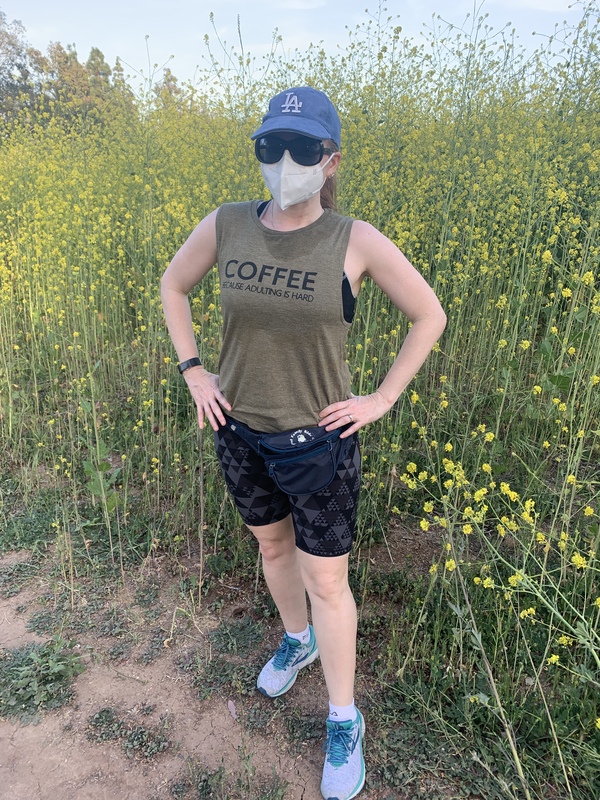This is a picture taken of a woman outdoors, who is wearing a face mask, sun glasses, a blue Los Angeles hat, a fanny pack, a tank top which reads 'Coffee: because adulting is hard", and dark shorts. 