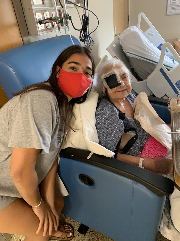Masked young woman poses with elderly woman sitting in a recliner.  The seated woman has an oxygen tank and a patch over one eye.