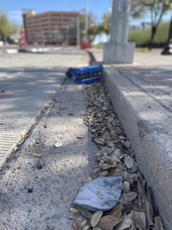 This is a picture of a face mask that has been discarded in a gutter on a street. Buildings can be seen in the blurry background. 
