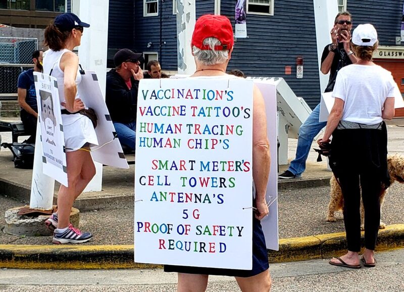 This is a picture of a man wearing  a sign protesting COVID-19 vaccinations. His sign reads "Vaccination's, Vaccine Tattoo's, Human Tracing, Human Chip's, Smart Meter's, Cell Towers, Antenna's, 5G- Proof of Safety Required."