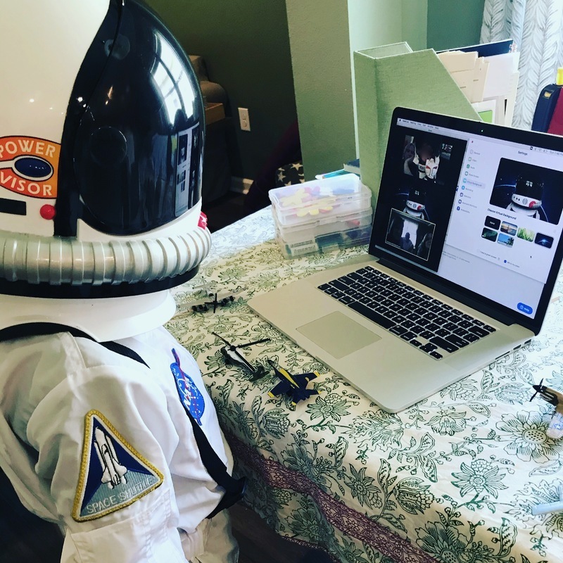 This is a picture taken of a person who is dressed in astronaut gear, and sitting at a table looking at a zoom call on a laptop.  
