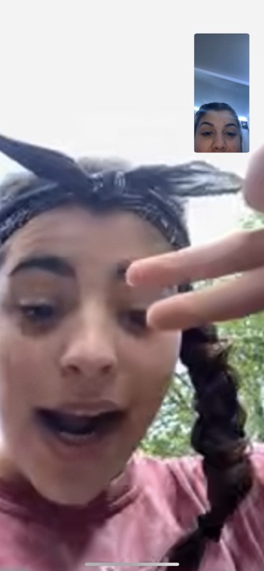 Video call screenshot of two females. One on the main screen has a bandana and braids making a peace sign. The other in the smaller screen to the top right only shows their nose and above of their face. 