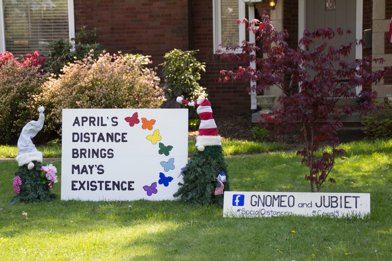 Photo of someone's front yard featuring two gnomes and a sign that reads "April's distance brings May's existence". 
