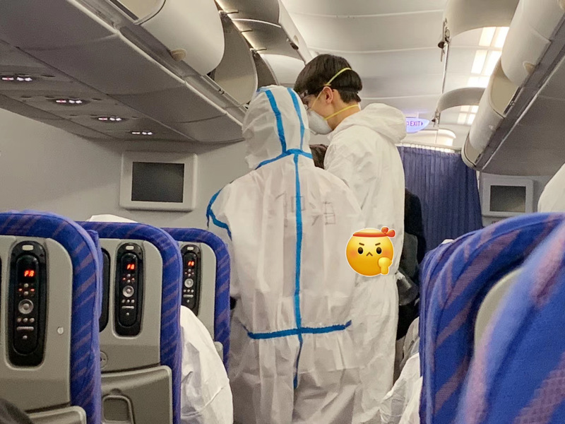 This is a picture taken of two people on a plane, wearing full body protective suits and face masks. 