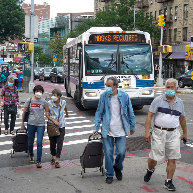 This is a picture of a group of people wearing face masks using a crosswalk to cross a busy street. A bus behind them has a message displayed on it reading: "Masks Required". 