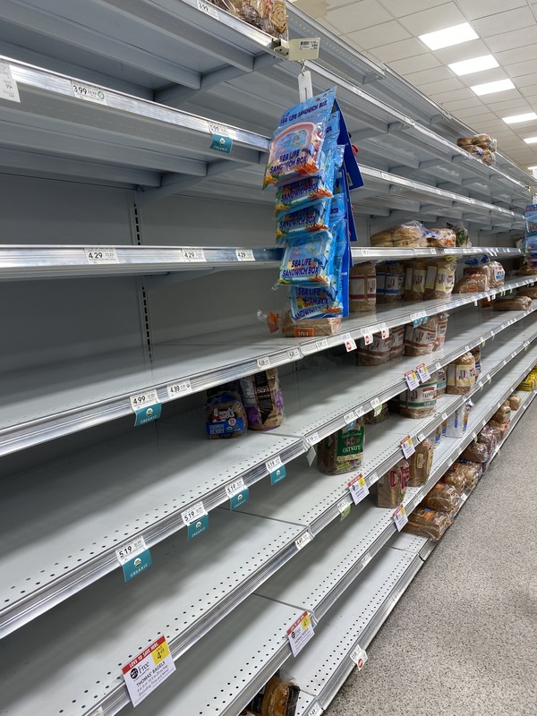 A bread aisle in a grocery store with a few loaves of bread left on the shelves. 