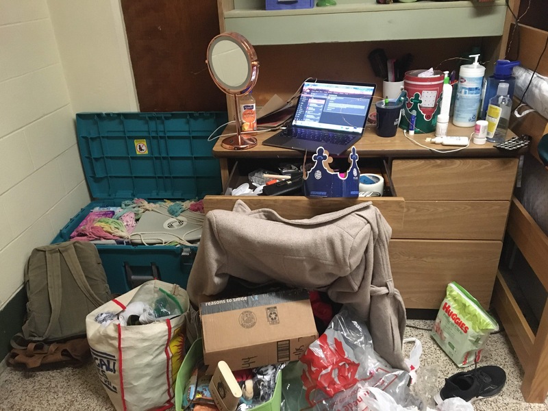 This is a picture taken of someone's messy dresser area. Multiple bags of other objects are scattered on the floor in front of the dresser. 