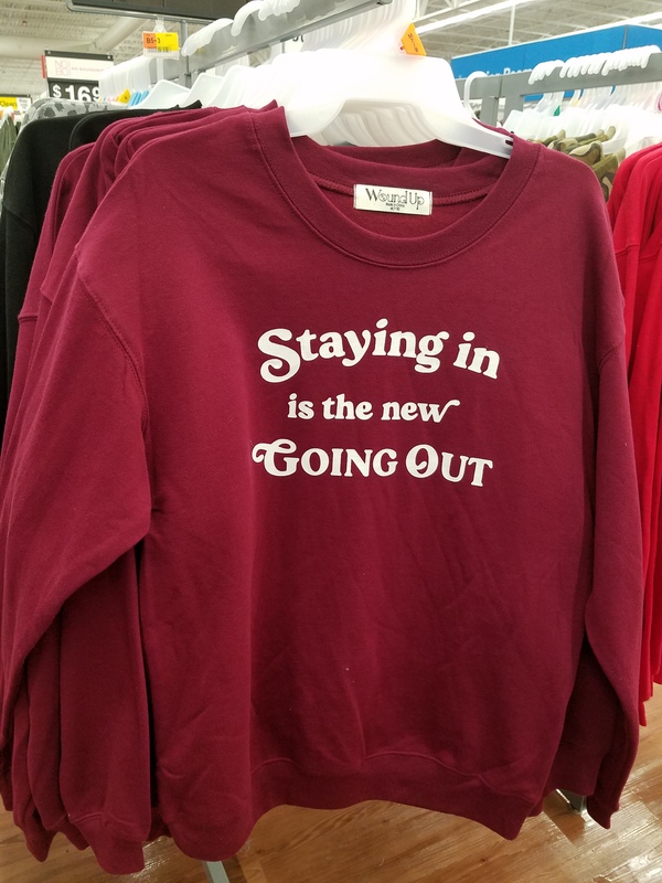 This is a picture of a red long sleeve shirt, which has the message "Staying in is the new going out" printed in white lettering on it. 