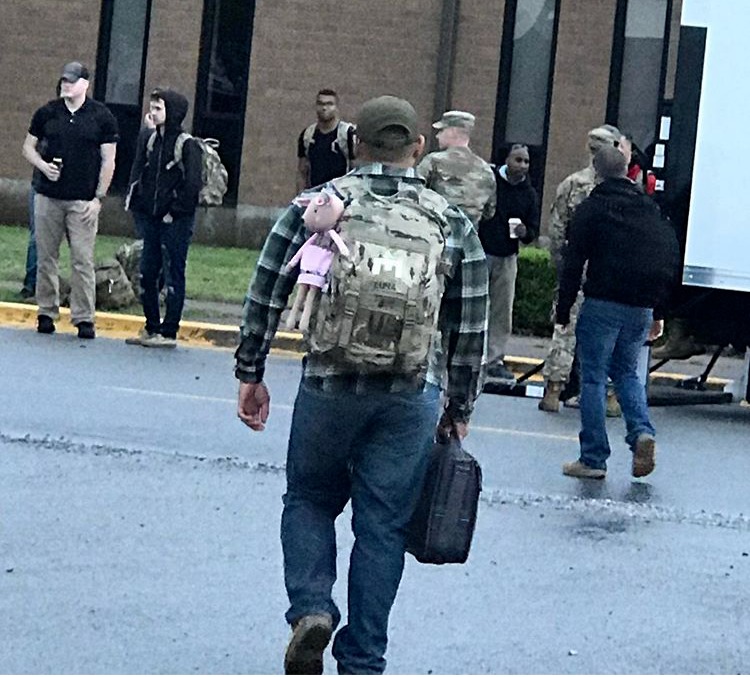Photo of a man wearing a military backpack with a child's stuffed animal attached standing in front of a school with other men in military backpacks and full camo uniforms. 