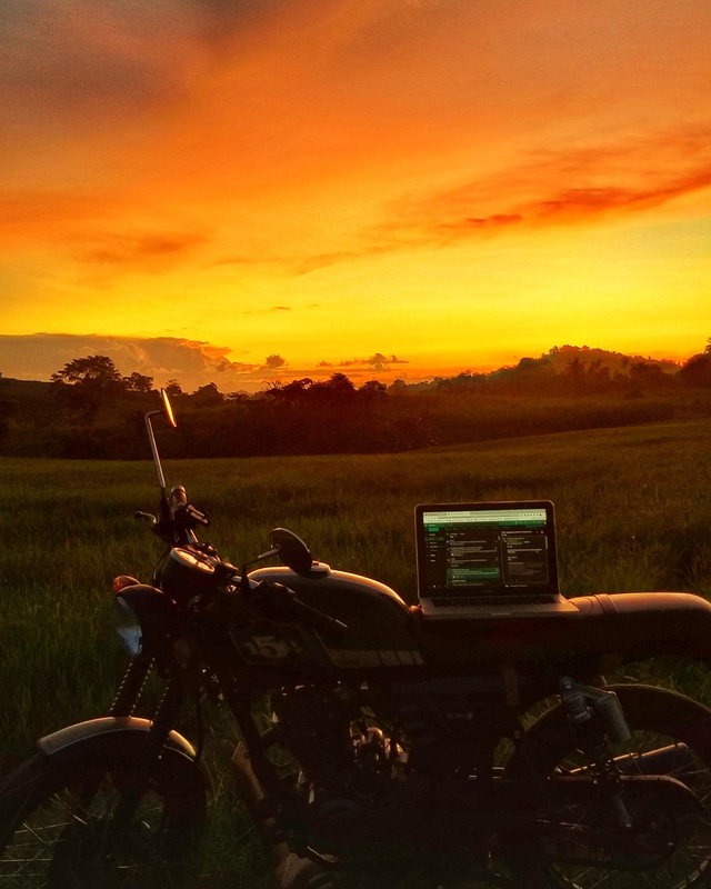 A photograph of a motorcycle with a laptop on top of it in front of a sunset. 