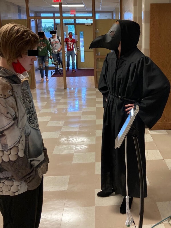 This is a picture of a person wearing a plague doctors mask and robe standing in a school hallway in front of some students. The students are wearing face masks. 