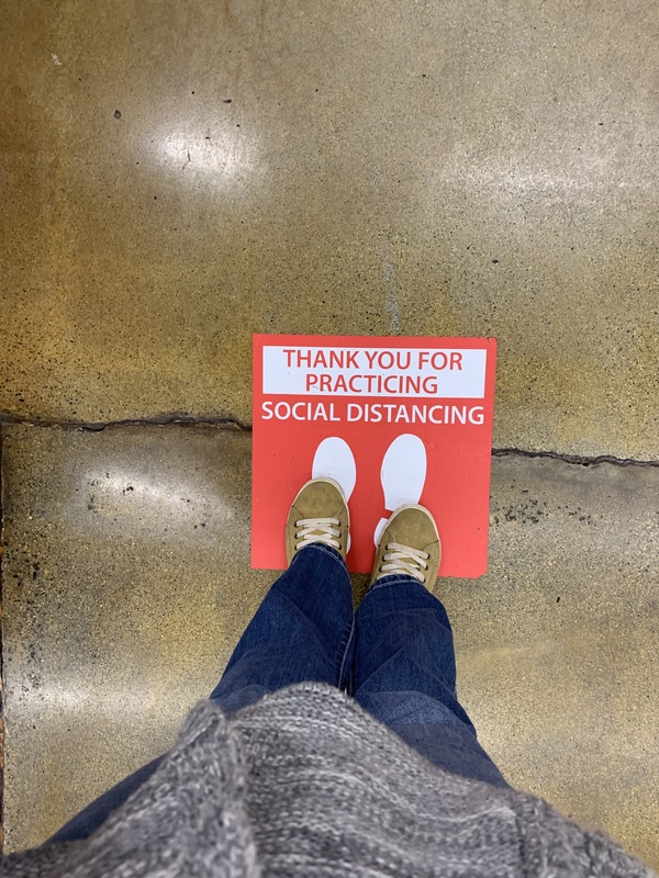 Image of someone's shoes, standing on top of a social distancing sign on the floor.