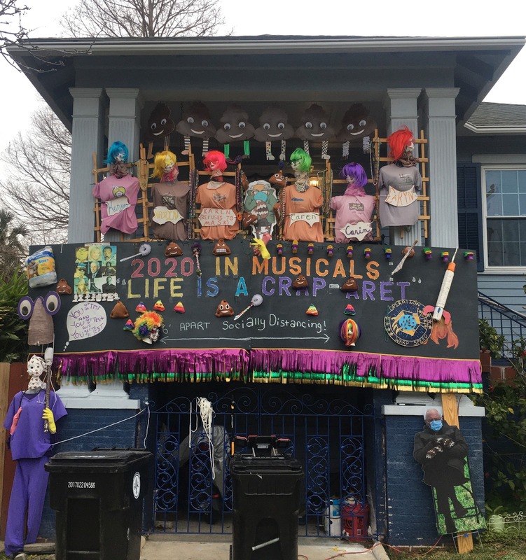 This is a picture taken of a person's house which is decorated with various items related to the COVID-19 Pandemic. A line of poop shaped emojis with smiley faces hang from the roof, under which sits a line of dummies with skulls for heads each labeled under a different spelling of the name "Karen", with a sign underneath stating that each dummy did not wear their mask. A banner underneath these objects reads "2020 in musicals: 'Life is a Crap-aret'". The bottom of the banner encourages the reader to follow social distance protocols. Several mannequins dressed in medial gear or face masks flank the banner. 