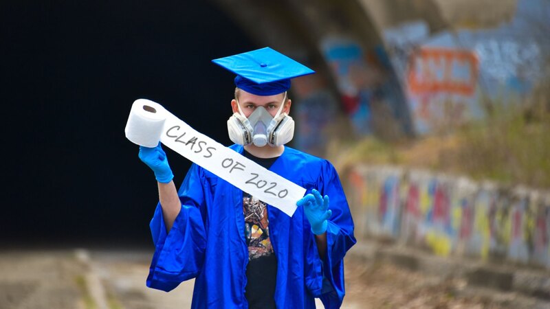 A graduating senior wearing a surgical face mask and gloves holding a roll of toilet paper reading "Class of 2020."