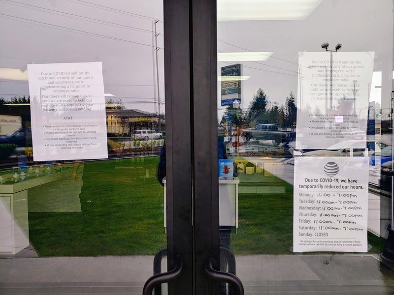 Two doors in front of a store have multiple papers taped on the inside of the glass informing patrons of their changed store hours and the decrease in people in the lobby. 