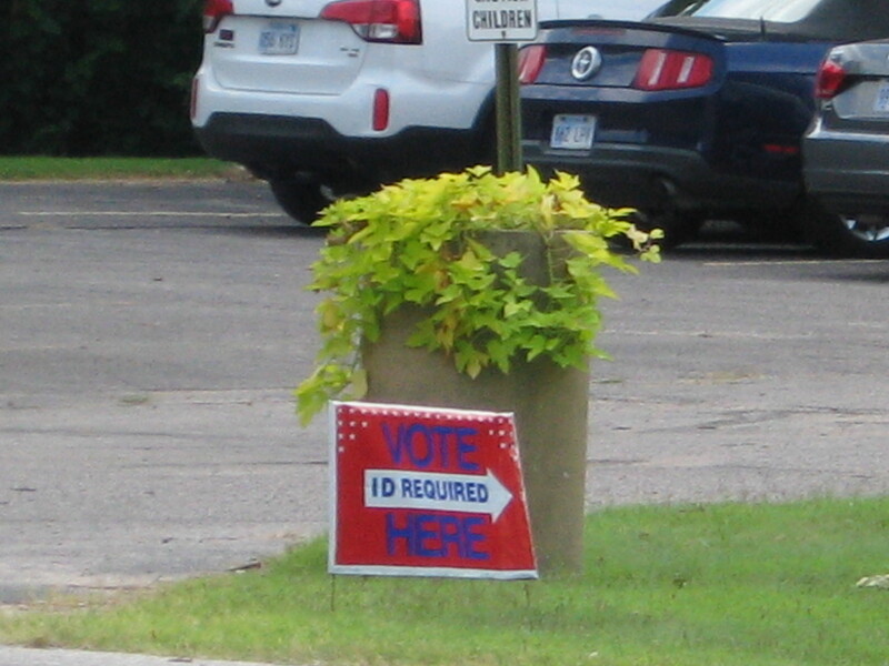 A red yard sign reading "VOTE HERE. ID required".