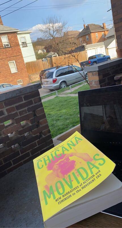 This is a picture taken of a book resting on a laptop which is sitting on a persons lap on a front porch. The book title reads "Chicana Movidas".