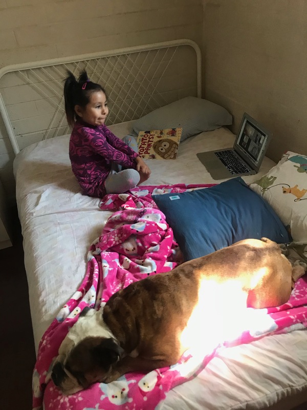 Picture of a young child sitting on a bed looking at a Zoom call on a computer.