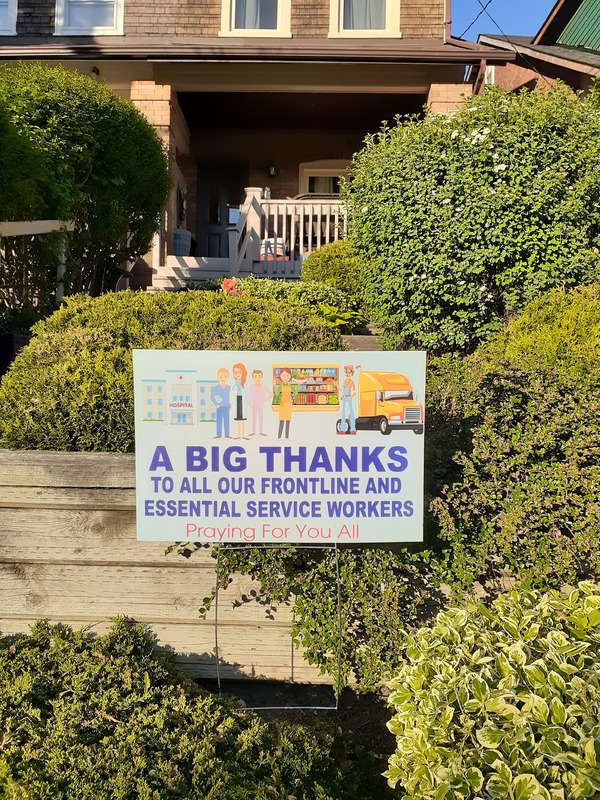 This is a picture of a sign posted in front of a home which reads "A big thanks to all our front line and essential service workers, praying for you all" along with pictures of caricatures of various essential workers. 