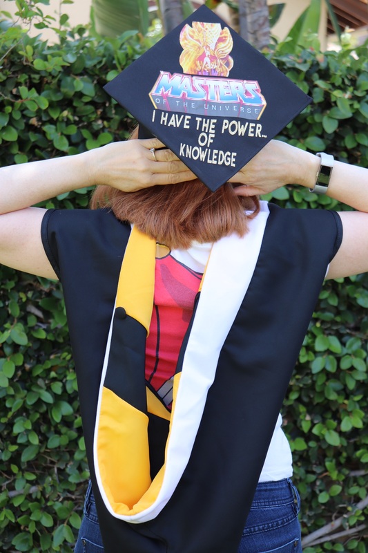 This is a picture taken of a women in graduation clothing, with her back turned to the camera. Her graduation cap is decorated with a message reading: "Masters of the universe, I have the power... of knowledge." 