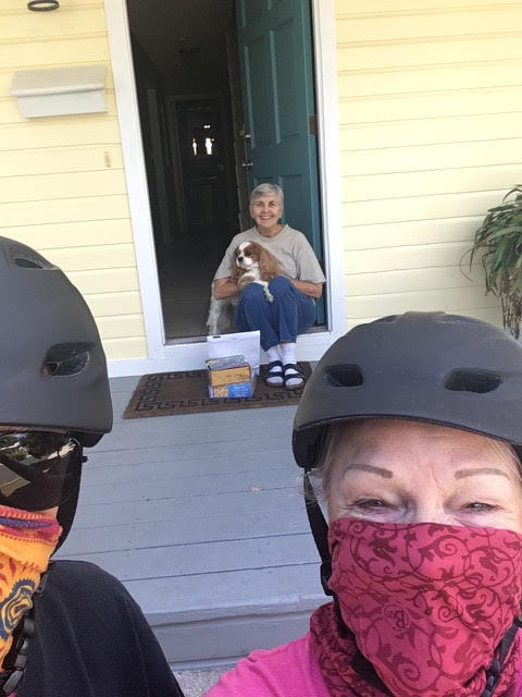 This is a picture of two people visiting another at the door to her house while socially distancing. The visitors wear bandanas and helmets, while the person who is being visited holds her dog on the front step of her house.