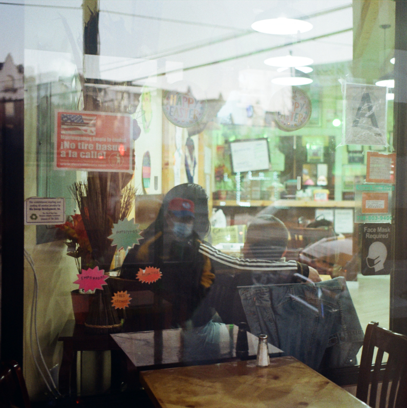 This is a picture taken from outside the front window of a business, showing two patrons wearing masks sitting at a table. Among the various signs posted on the window, one reads "face mask required". 