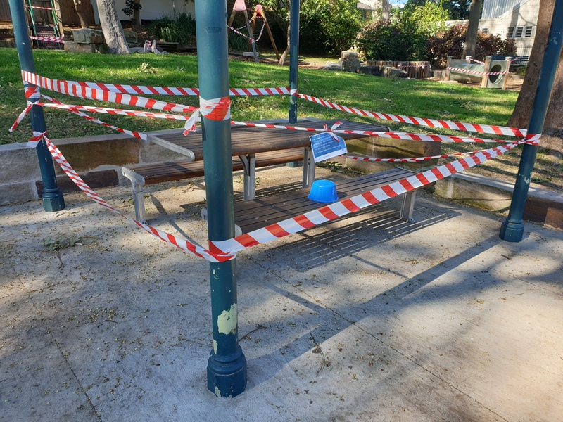 Photo of a park. All seating areas and playground equipment are blocked off with ted and white tape.