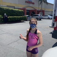 This is a picture of a young girl wearing a gymnastics uniform and a face mask, standing in front of a gymnastics building. She is giving a thumbs up sign.