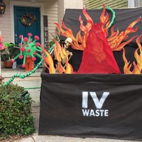 This is a picture taken of a house decorated in items related to COVID-19. A depiction of a COVID-19 germ sits on the front steps, and a fake dumpster with a skeleton and flames inside of it sits in front of the steps. The dumpster is labeled "IV Waste."