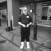 A man is wearing white rain boots with pants tucked into the boots. He is wearing a polo shirt with a name tag attached to it. The man is standing in front of a grocery store. The man is wearing circular sunglasses. 