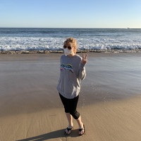 This is a picture of a woman wearing a face mask and sunglasses at the beach, posing for a photo by making a peace sign with one hand. 