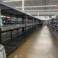 A supermarket is shown with white walls and brown concrete flooring. The empty isle has black and grey shelves on the right and on the left is white shelves that has various cleaning supplies. 