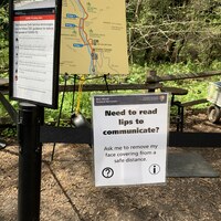This is a picture of a sign at the start of a trail, it reads: "Need to read lips to communicate? Ask me to remove my face covering from a safe distance." A map, and information about the National Park Service is posted next to this sign.